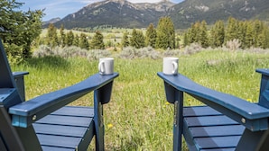 Two Adirondack chairs with white mugs on them, set against a backdrop of a grassy field and mountains. - Ski. Hike. Soak. Dogs Welcome | Nordic Ski Haus (Big Sky)