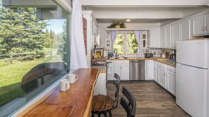 A kitchen with white cabinets, a wooden countertop, and a view of a green landscape. - Ski. Hike. Soak. Dogs Welcome | Nordic Ski Haus (Big Sky)