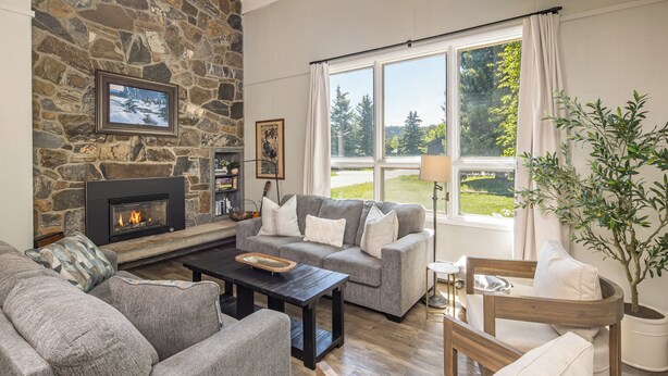 A living room with a stone fireplace, grey sofas, a wooden coffee table, and a view of a green landscape through large windows. - Ski. Hike. Soak. Dogs Welcome | Nordic Ski Haus (Big Sky)