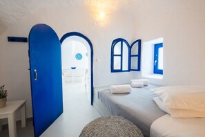 A room with a blue door, a bed with white linens, a small table, and a wall-mounted shelf.