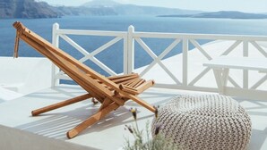 A white outdoor seating area with a wooden lounge chair and a knitted pouf, overlooking a scenic view of the sea and distant mountains.