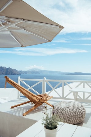 A white outdoor seating area with a wooden lounge chair and a knitted pouf, overlooking a scenic view of the sea and distant mountains. - Blu Bianco cave loft (Oia)