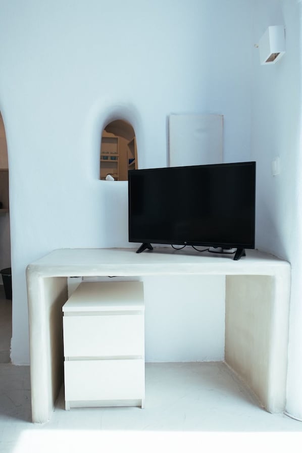 A white desk with a flat-screen TV, a small drawer, and a white wall with a light switch and a mirror.