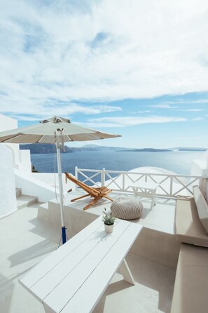 A white outdoor seating area with a view of the sea, a wooden lounge chair, a white sofa, and a small potted plant.