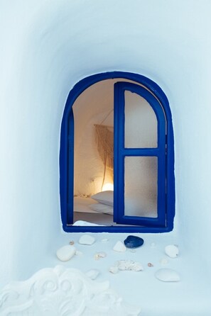A small, arched window with a blue frame and frosted glass panes, revealing a bed with white bedding and a hanging light fixture inside a room with a white, textured wall. - Blu Bianco cave loft (Oia)