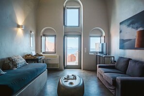 Un salon moderne avec un canapé, un pouf et un meuble pour la télévision. Il y a des fenêtres en arc de cercle et une vue sur l’extérieur.