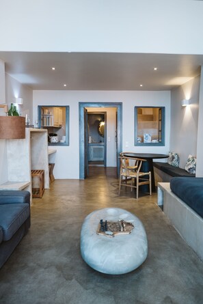 Un salon moderne avec un pouf rond, un canapé bleu, une table à manger avec des chaises, et un coin cuisine à l’arrière-plan.