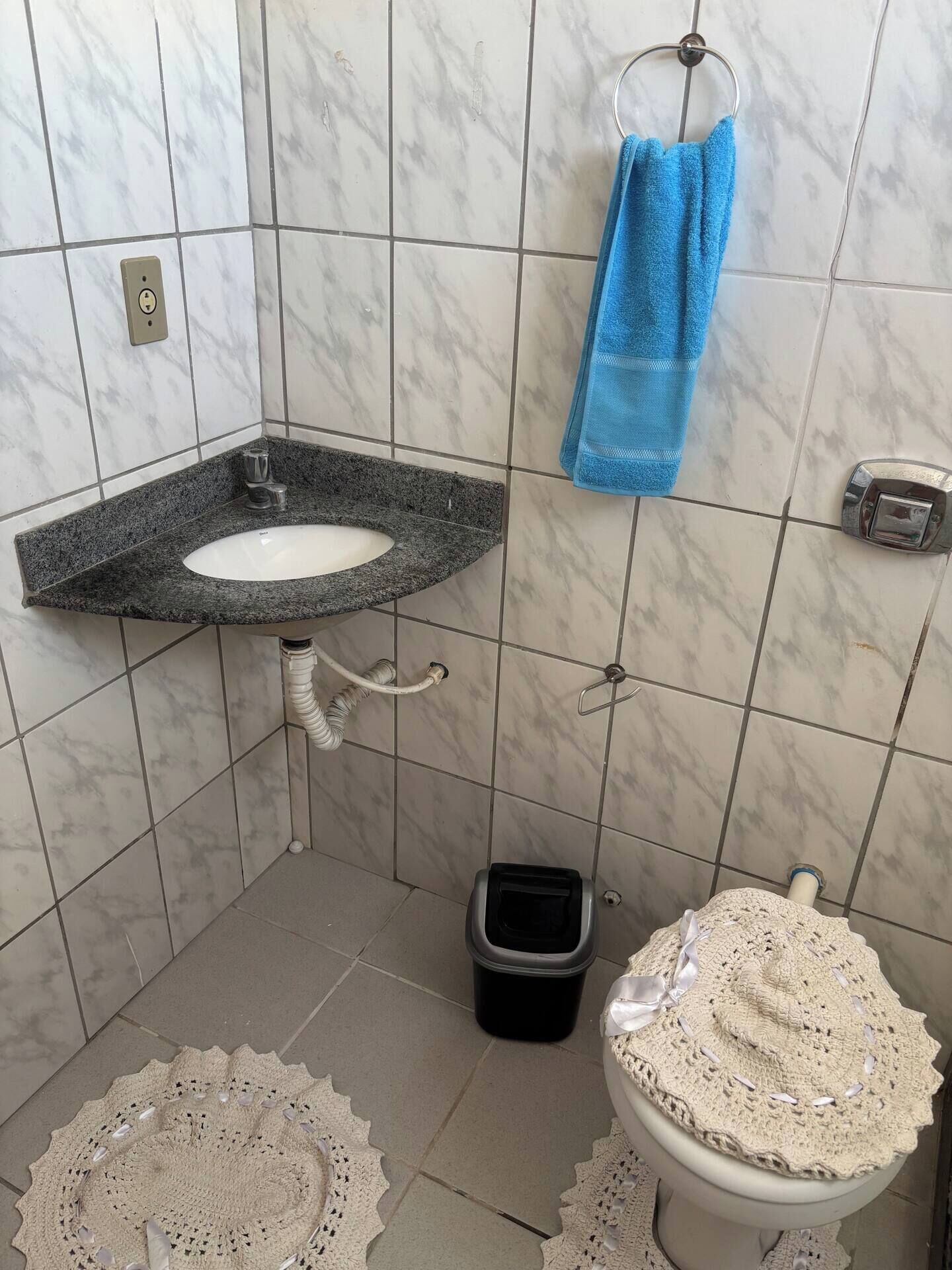 Un baño con pared de azulejos de mármol, un lavamanos de granito, una toalla azul, un inodoro con cubierta de crochet y un cubo de basura negro.