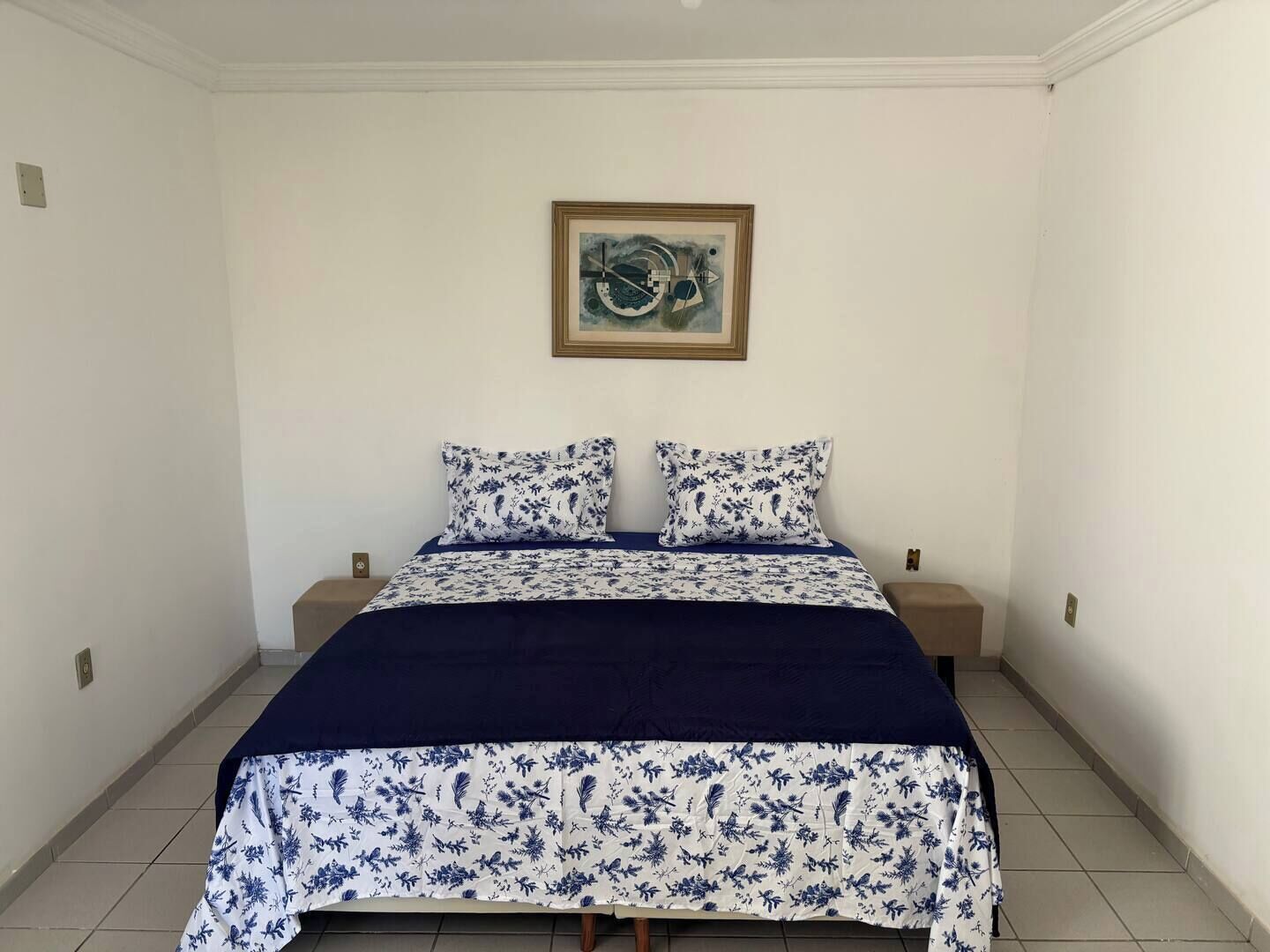 A bedroom with a bed, two pillows, a framed artwork on the wall, and a tiled floor.