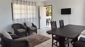 A dining area with a dark wooden table and chairs, a white door leading outside, and a flat-screen TV mounted on the wall. - Blue Wave Hostel (Serra)