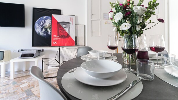 Un comedor moderno con una mesa lista para dos, con platos blancos, vasos transparentes y un florero con flores.