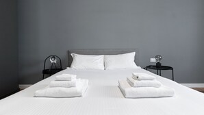 Una cama bien hecha con sábanas y toallas blancas, un cabecero gris y dos mesitas de noche con lámparas y un globo terráqueo.