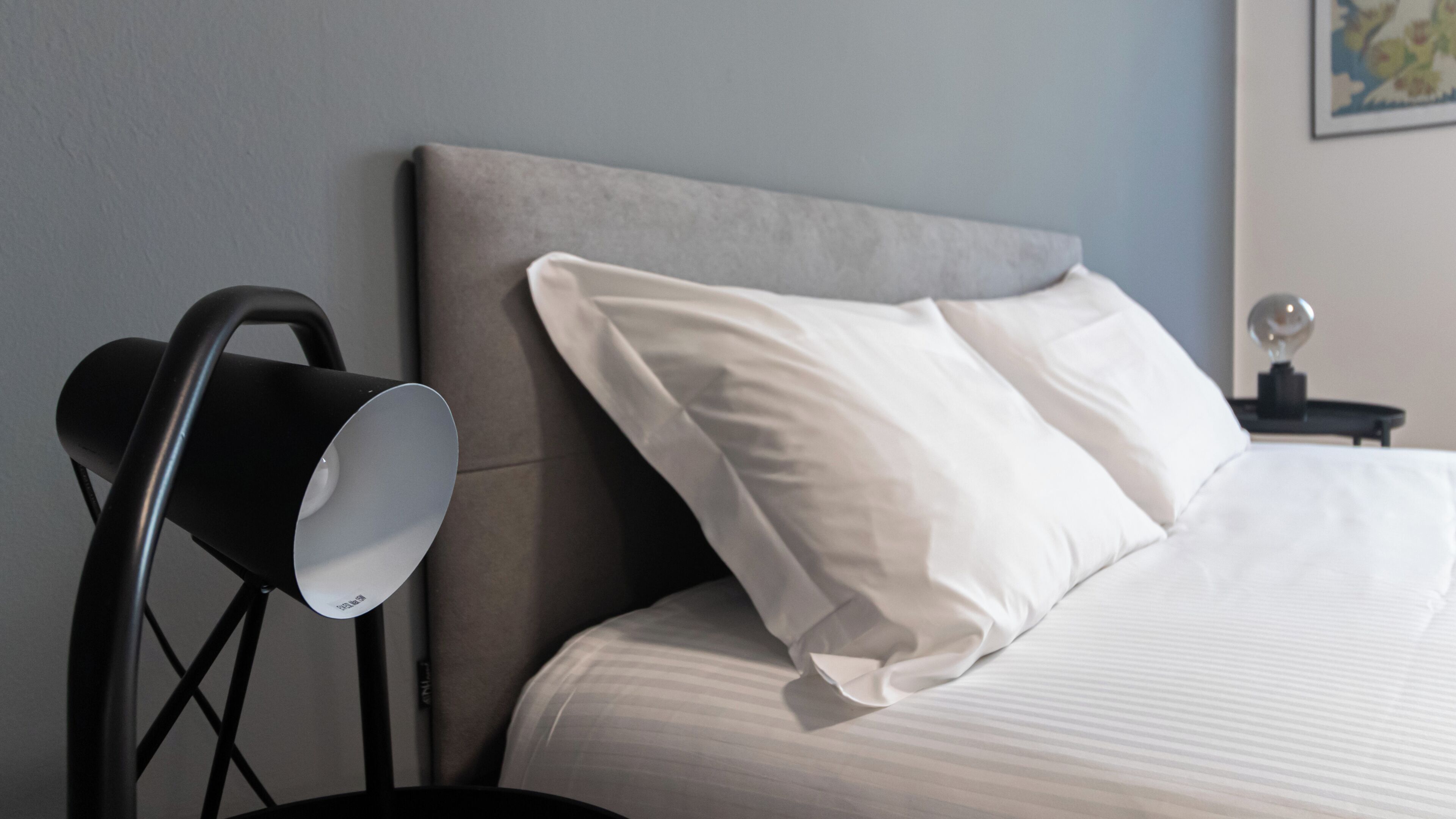 Un lit bien fait, avec du linge de lit blanc, une tête de lit grise et une lampe de sol noire.