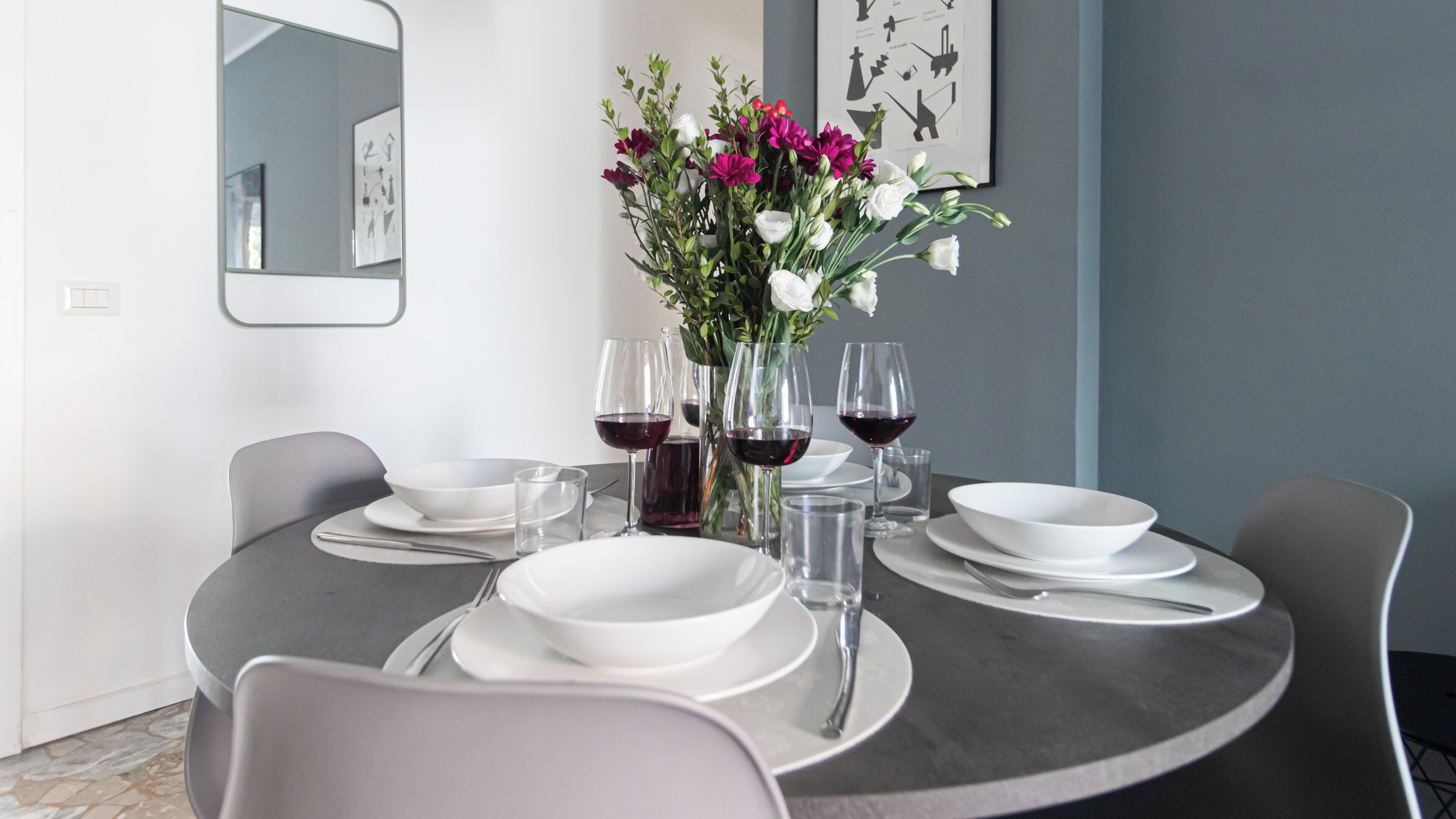 A round dining table with white plates, bowls, and cutlery, accompanied by wine glasses and a vase of flowers.