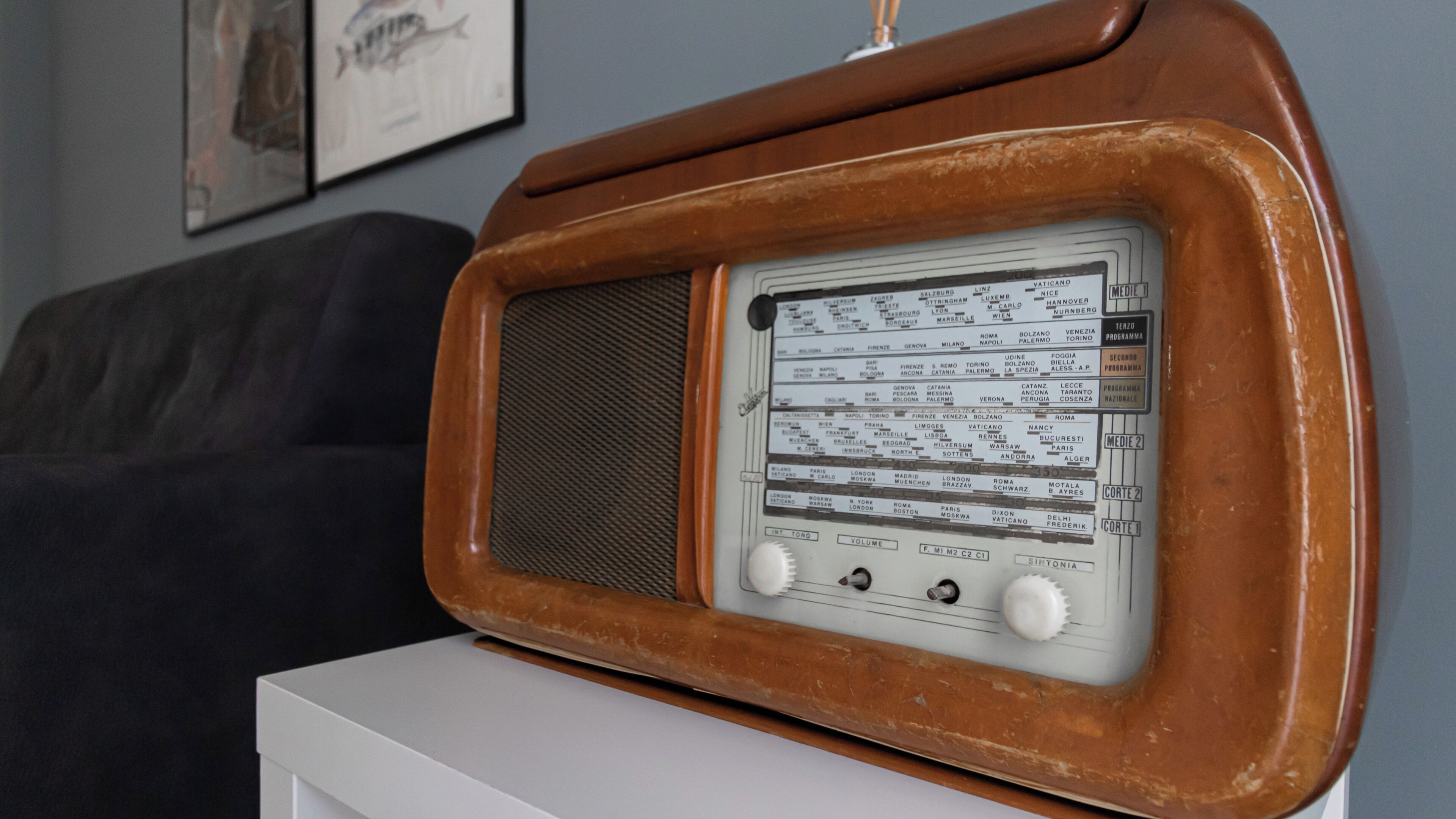 A vintage radio with a wooden frame and a white base, placed on a white table in a room with a dark-colored sofa and framed pictures on the wall.