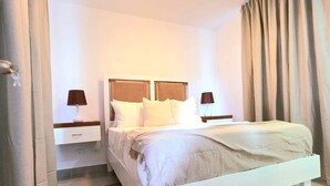 Une chambre à coucher moderne avec un lit blanc, des rideaux beiges et un sol carrelé.