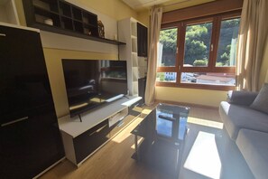 A modern living room with a flat-screen TV, a glass coffee table, a sofa, and a large window with a view of greenery. - 21A07 Apartment in Felechosa by R2R Consulting (Aller)