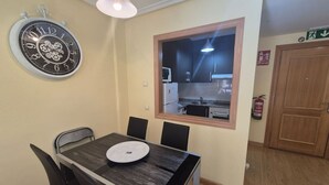 A dining area with a table, chairs, a wall clock, a window with a view of a kitchen, and an exit sign. - 21A07 Apartment in Felechosa by R2R Consulting (Aller)
