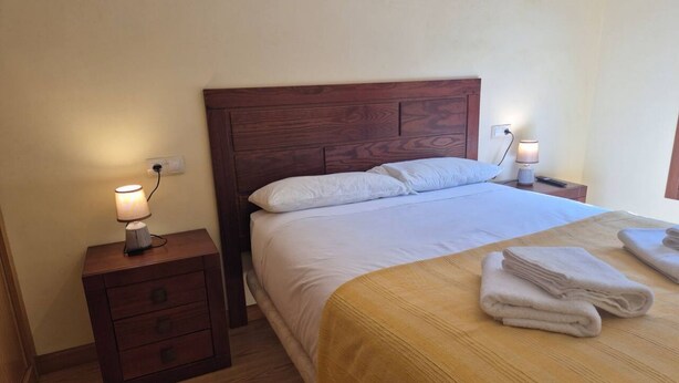A neatly made bed with a wooden headboard, a bedside table with a lamp, and a folded towel on the floor. - 21A07 Apartment in Felechosa by R2R Consulting (Aller)