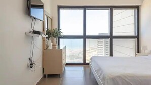 A modern bedroom with a large window, a bed, a dresser, and a flat-screen TV mounted on the wall. - Luxury Apt Tower Best Sea View Bat Yam (Bat Yam)