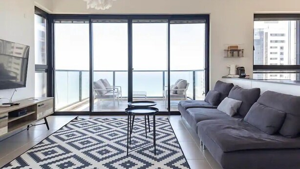 A modern living room with a grey sofa, a patterned rug, a TV, and a balcony with a view of the city. - Luxury Apt Tower Best Sea View Bat Yam (Bat Yam)