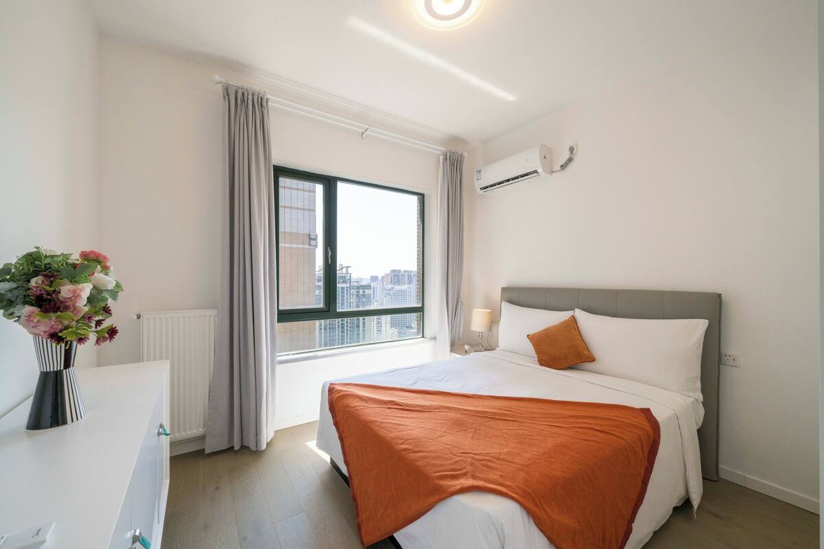 Een moderne hotelkamer met een groot bed, een nachtkastje, een bloemenvaas, een raam met gordijnen en een airconditioningsunit.