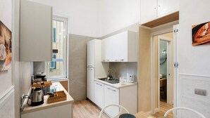 A compact kitchen with white cabinets, a coffee machine, and a dining area with a table and chairs. - Merliani House Holiday Home (Napoli)
