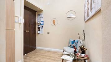 Un couloir avec une porte en bois, un tableau d’affichage, une table en verre avec des livres, une chaise et un tableau accroché au mur.