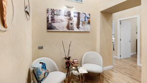 A room with two chairs, a small table with books and a vase, a large abstract painting, and a door leading to another room. - Merliani House Holiday Home (Napoli)
