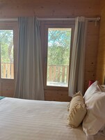 Habitación tipo cabaña de madera con una cama, cortinas y una ventana con vistas a árboles.