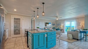 A spacious kitchen with a turquoise island, a dining area with a blue table, and a living room with a gray sofa and a ceiling fan. - Awesome Beachfront | Gulf Views + Pools + Tennis + Pickleball & Beach Access (Port Aransas)