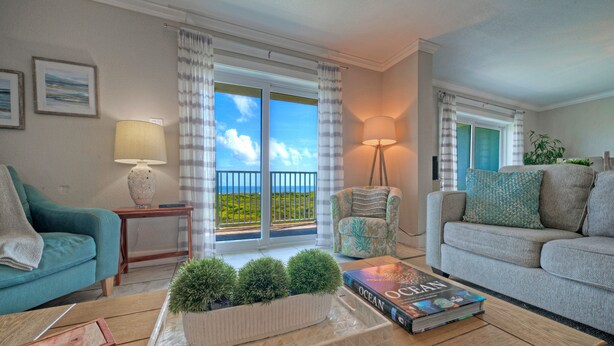 A living room with a sofa, a coffee table with a book, a lamp, and a view of the outdoors through a window. - Awesome Beachfront | Gulf Views + Pools + Tennis + Pickleball & Beach Access (Port Aransas)