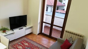 Ein kleiner Wohnraum mit einer Couch, einem Fernseher auf einem Schrank, einem roten Teppich mit Muster und Blick auf ein rotes Ziegeldach.
