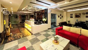 A hotel lobby with a reception desk, a red sofa, a coffee table with newspapers, and a checkered floor. - V Hotel TSSG (New Delhi)