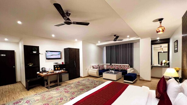 A hotel room with a bed, a sofa, a desk with a computer, a television, and a ceiling fan. - V Hotel TSSG (New Delhi)