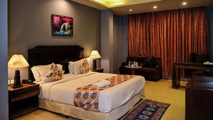 A hotel room with a bed, two bedside tables with lamps, a desk, a chair, a small table with bottles, and a large window with curtains. - V Hotel TSSG (New Delhi)