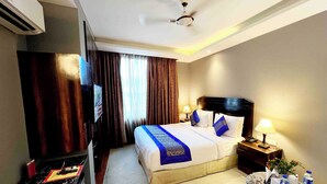 A hotel room with a bed, a TV, a desk, a lamp, and a ceiling fan. - V Hotel TSSG (New Delhi)