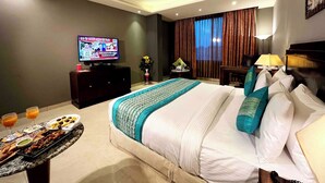 A hotel room with a bed, a television displaying a news channel, a dining table with food and drinks, and a ceiling fan. - V Hotel TSSG (New Delhi)