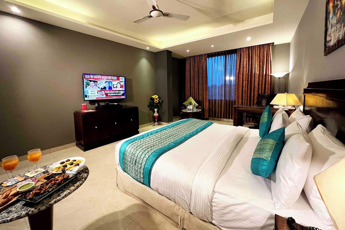 A hotel room with a bed, a television displaying a news channel, a dining table with food and drinks, and a ceiling fan.