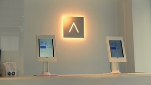Dos ordenadores Apple iMac sobre un escritorio blanco de diseño minimalista, un letrero iluminado montado en la pared y un altavoz pequeño a la izquierda.