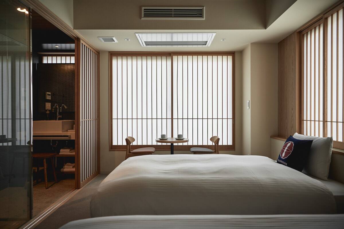 Een hotelkamer met een groot bed, een klein tafeltje met stoelen, een raam met verticale jaloezieën en een schuifdeur naar een badkamer.