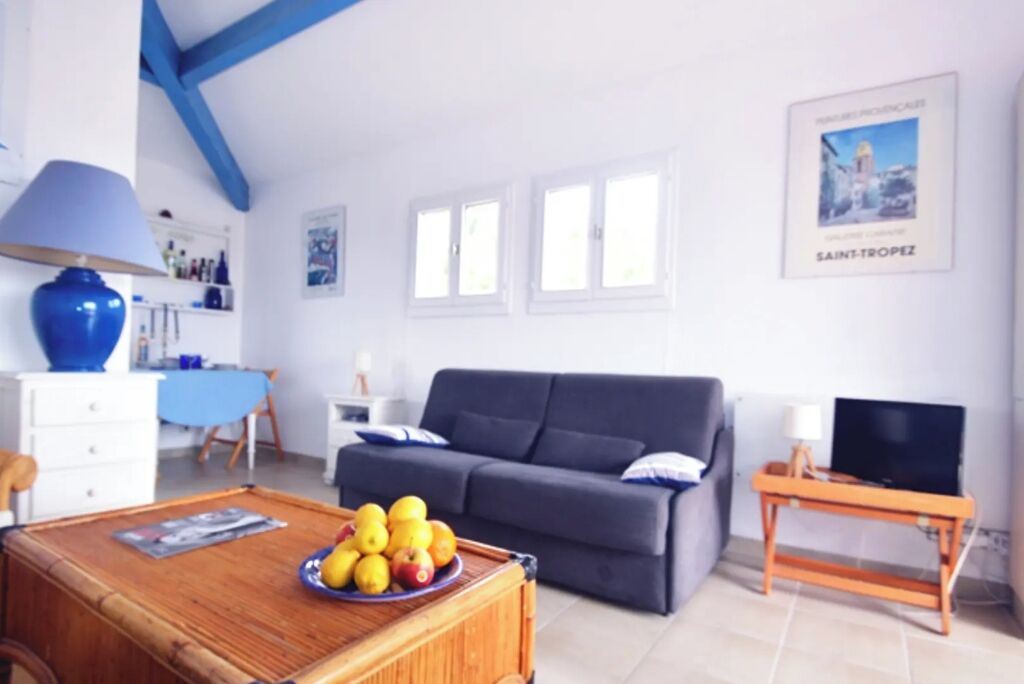 Un salon avec un canapé, une table basse sur laquelle on a posé des fruits, un téléviseur sur un meuble et un mur orné d’une affiche de Saint-Tropez.