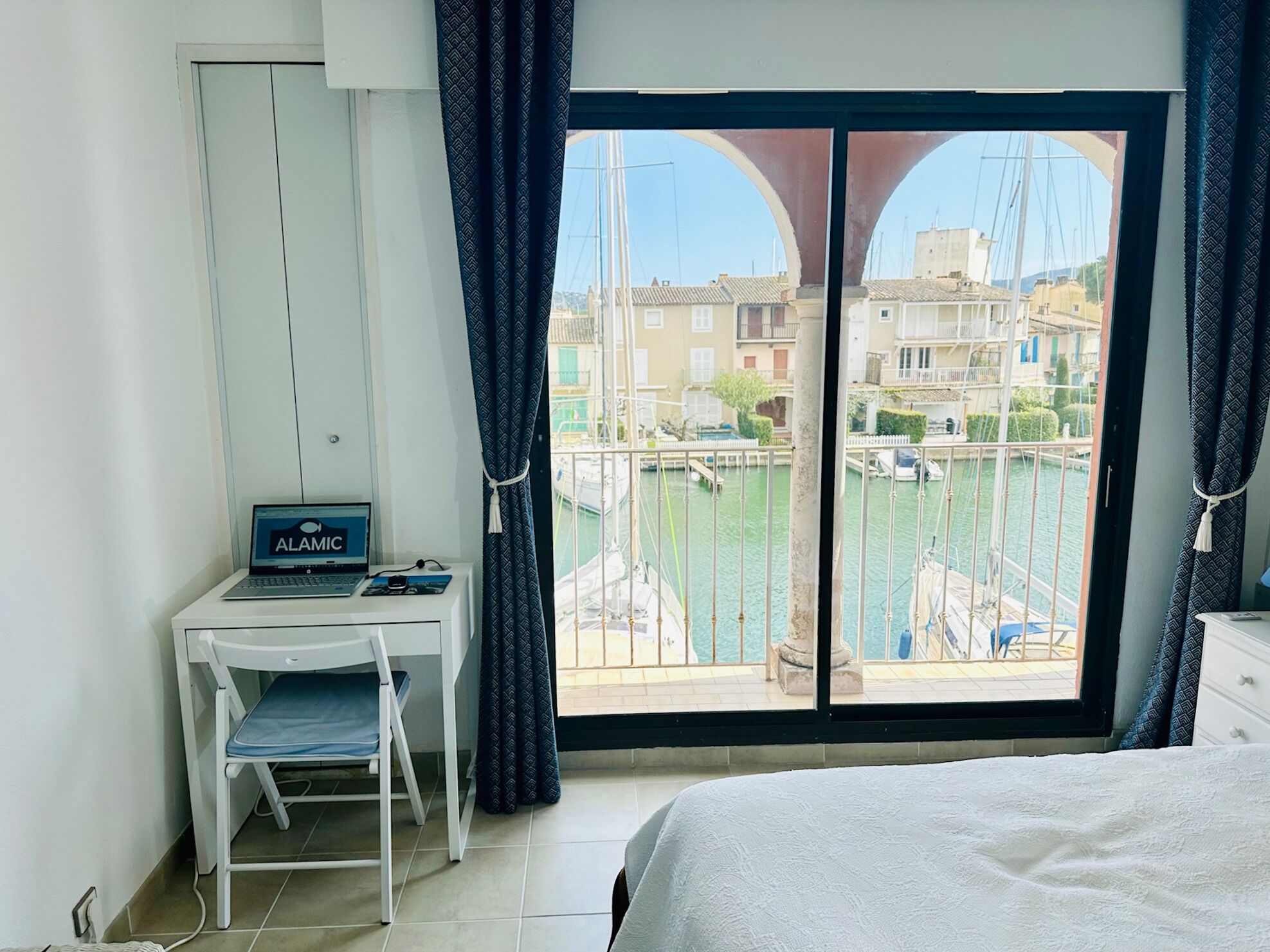 A room with a bed, a desk with a laptop, and a view of a marina with boats and buildings.