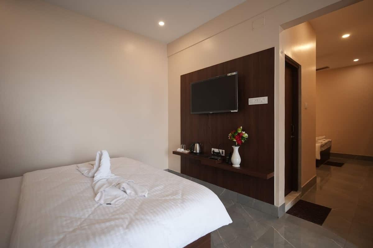 A hotel room with a bed, a TV mounted on the wall, a small desk with a vase of flowers, and a bathroom visible through an open door.