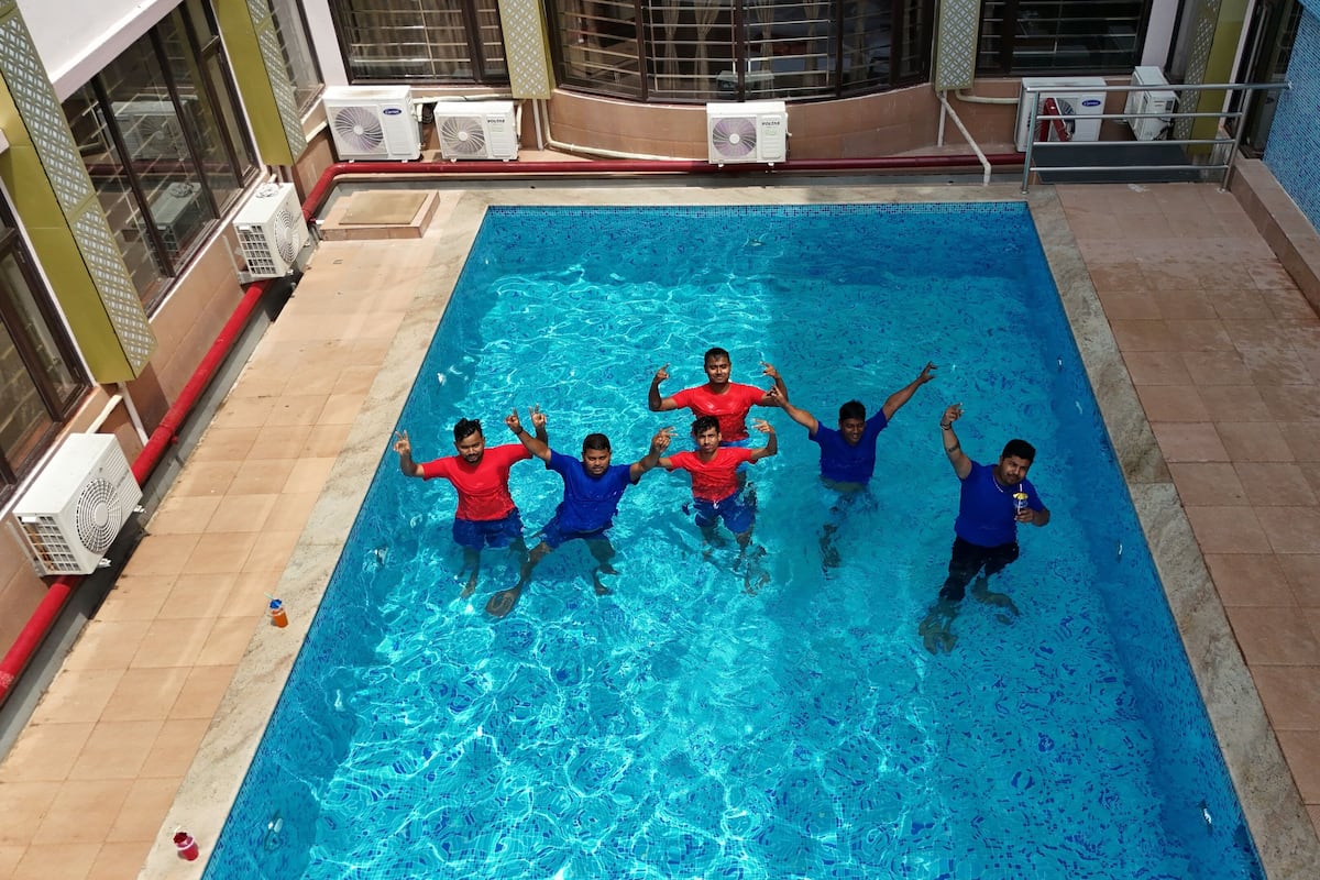 A swimming pool with four people in the water, surrounded by air conditioning units and a tiled wall.