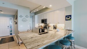 A modern kitchen with a marble countertop, white cabinets, and a stainless steel sink. There are three bar stools and a microwave above the counter. - 1002E-Beautiful 2 bedroom and 2 bath newly remodeled unit (Orange Beach)