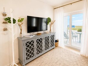 Ein Wohnzimmer mit Flachbildfernseher, einer grauen Kommode mit Zieraussparungen und einem Balkon mit Sitzgelegenheit.
