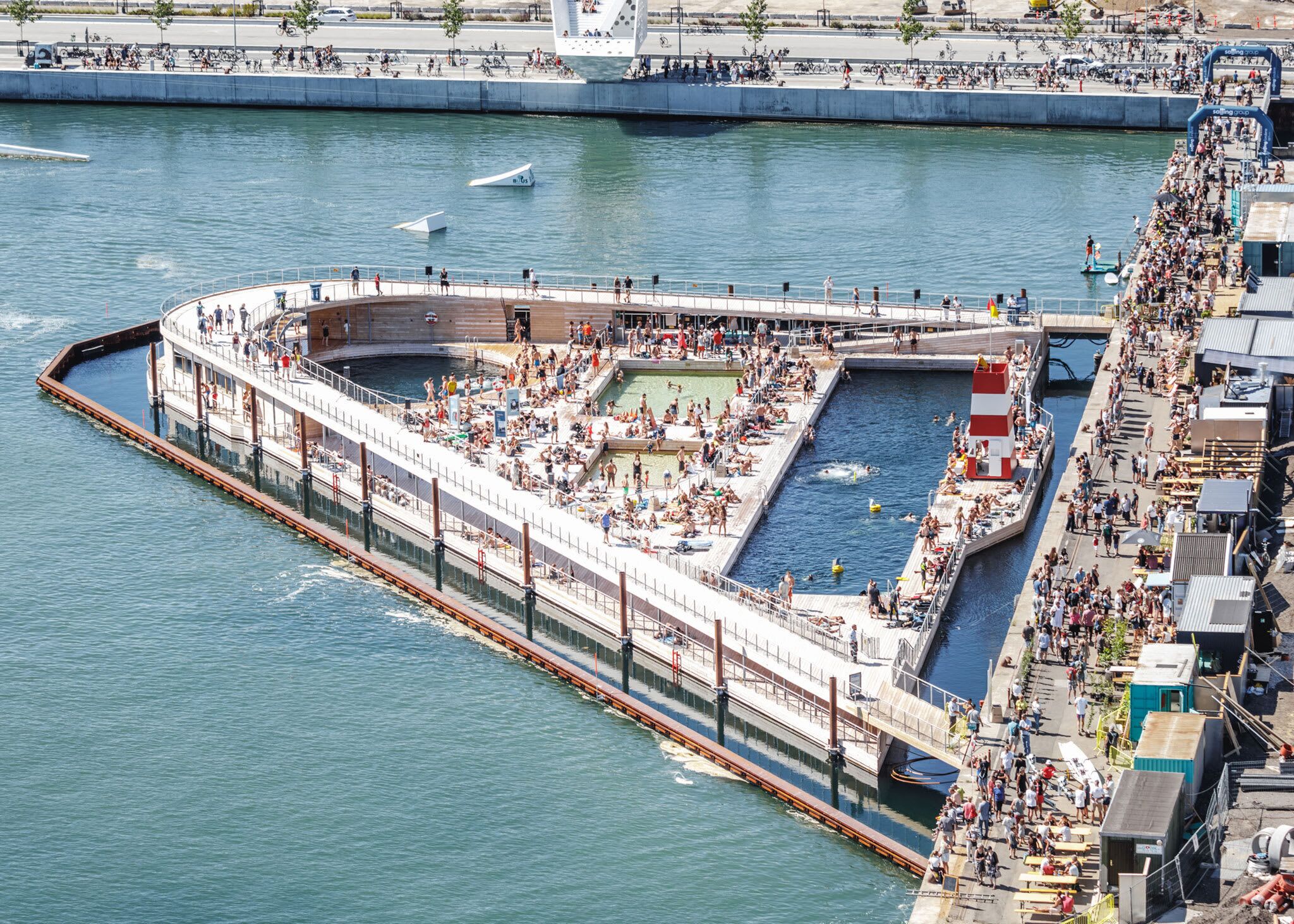 A waterfront swimming pool surrounded by a high fence, with people swimming and sunbathing.