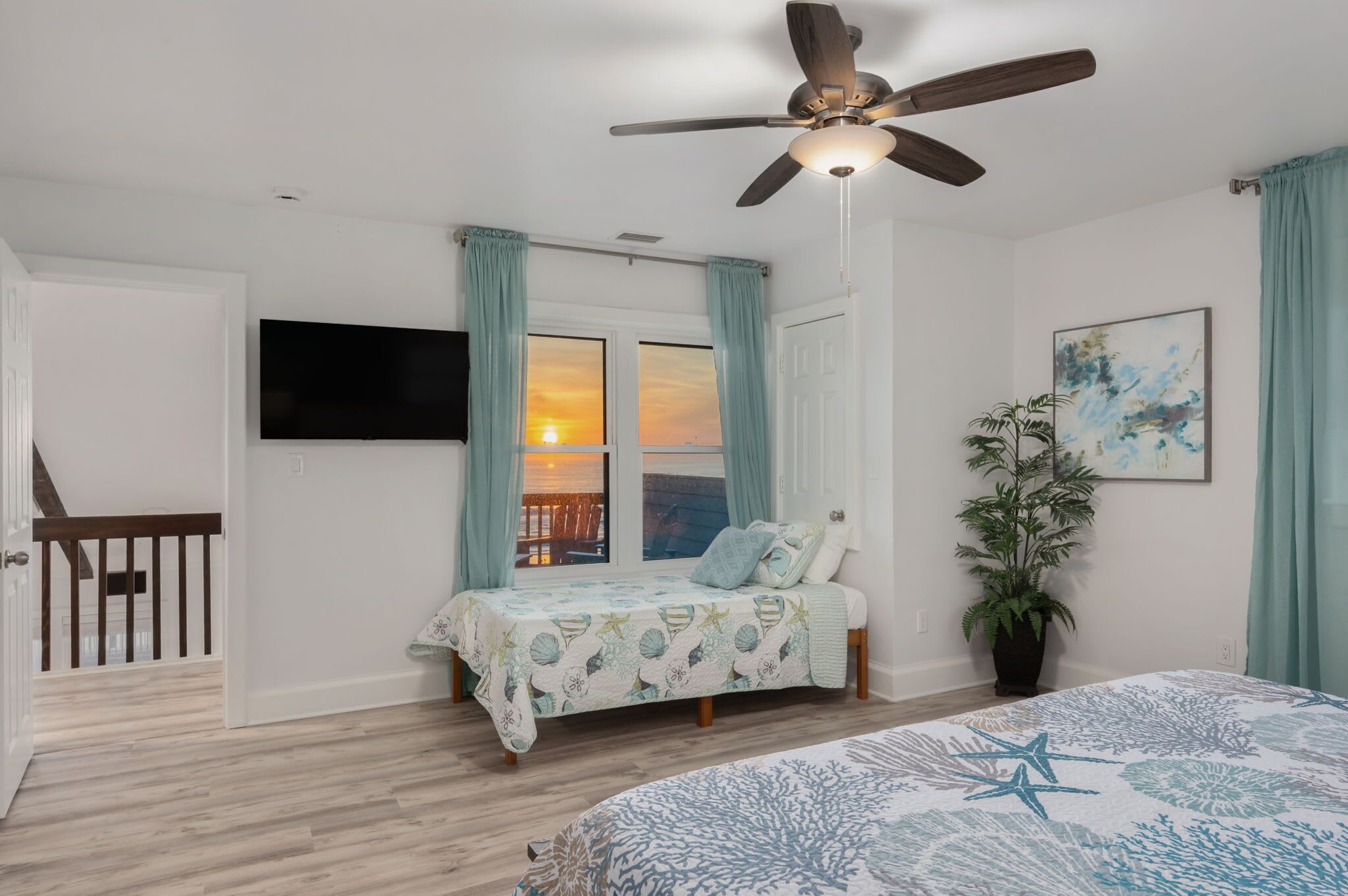 A bedroom with a bed, a ceiling fan, a window with curtains, a plant, and a painting on the wall.