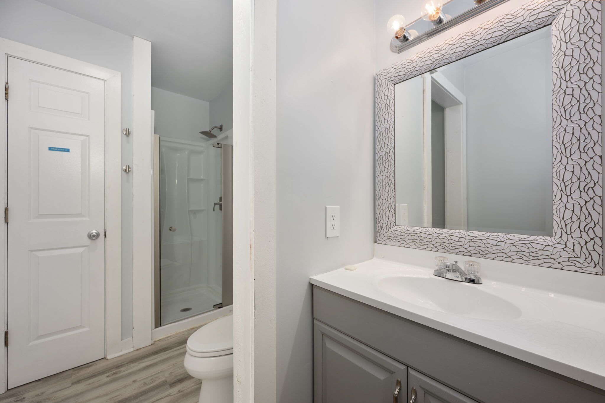A bathroom with a white door, a shower, a sink with a mirror, and a toilet.